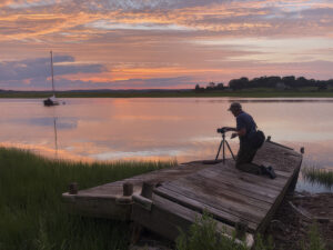 image of phil nelson kneeling on a dock at sunset taking a picture of the sunset